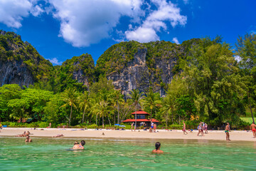 Bungalow house with red roof among coconut palms near the cliffs with people sunbathing and swimming in emerald water on Railay beach west, Ao Nang, Krabi, Thailand