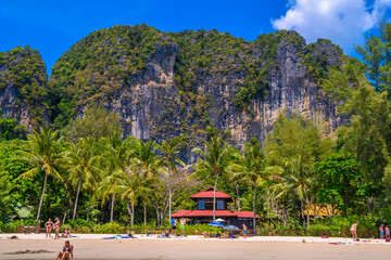 Bungalow house with red roof among coconut palms near the cliffs with people sunbathing and swimming in emerald water on Railay beach west, Ao Nang, Krabi, Thailand