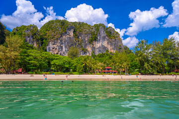Obraz premium Bungalow house with red roof among coconut palms near the cliffs with people sunbathing and swimming in emerald water on Railay beach west, Ao Nang, Krabi, Thailand