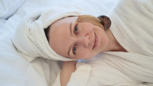 Top view of beautiful woman with bath towel on head holding cute snail on face while lying on soft blanket indoors. Peaceful young adult getting cosmetology treatment by using healing mucus on skin.