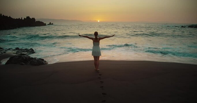 Back view of relaxed barefoot woman with outstretched arms on sandy beach at sunset.