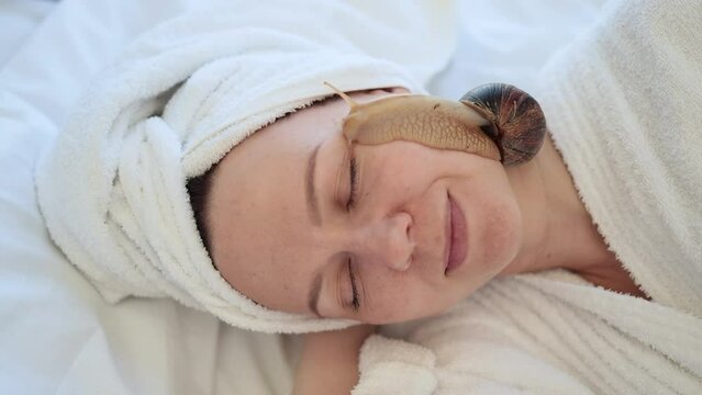 Top view of beautiful woman with bath towel on head holding cute snail on face while lying on soft blanket indoors. Peaceful young adult getting cosmetology treatment by using healing mucus on skin.
