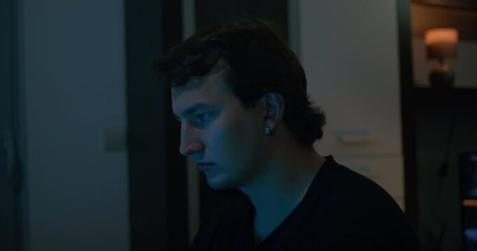Young man with earrings sits in dark and empty room searching Internet browser on laptop. Guy face glowing from light of laptop screen