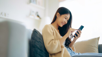 Happy young asian woman relax on comfortable couch at home texting messaging on smartphone, smiling girl use cellphone, chatting online message, shopping online from home - Powered by Adobe