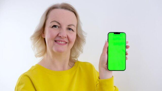 Smiling mature woman posing holding phone with tracking points on blank empty green screen, isolated on orange background. Giving ILY I Love You hand sign. High quality 4k footage