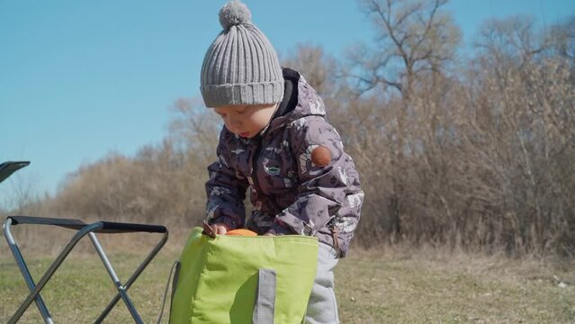 A boy in warm casual clothes takes out a camping coocking pot, a child helps his parents cook food while hiking. Family holidays with children. Toddler learns to set up a campsite while on an journey.