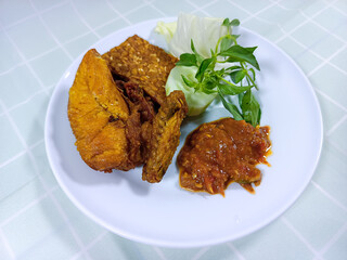 Close-up view of Ayam Penyet on a white plate. An Indonesian dish consists of fried chicken and tempeh, served with spicy sambal, a slice of cucumber, cabbage, and lemon basil. Indonesian street food.