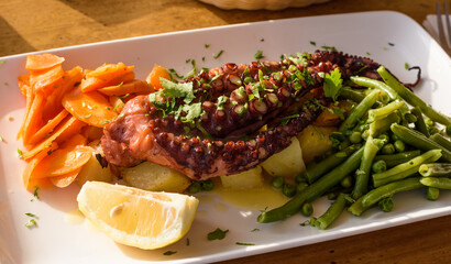 Delicious grilled octopus tentacles with healthy and fresh vegetables, potatoes, lemon slice and string beans with green peas and carrots on a white plate. Healthy seafood