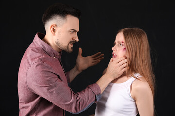 Angry young man threatening his bruised wife on dark background. Domestic violence concept