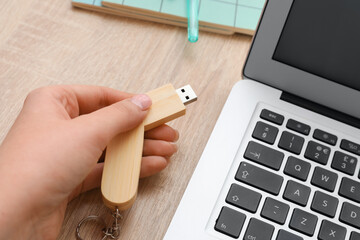 Female hand with USB flash drive and laptop on wooden background, closeup