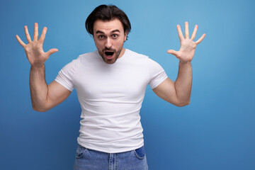 brutal young brunette guy in a white t-shirt scares on a studio background