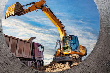 A wheeled excavator loads a dump truck with soil and sand. An excavator with a high-raised bucket against a cloudy sky View from the trench. Removal of soil from a construction site or quarry.