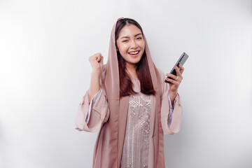 Fototapeta premium A young Asian Muslim woman with a happy successful expression wearing a hijab and holding smartphone isolated by white background