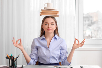 Young businesswoman with cup and books on her head meditating in office. Balance concept