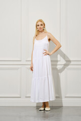 Fototapeta premium A beautiful woman with long hair in a white sundress stands indoors and looks into the camera