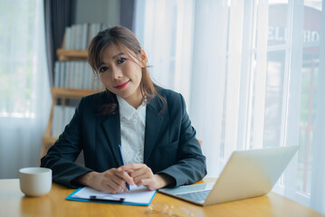 Beautiful asian business woman looking at camera in her office by her table there are important documents, a laptop and a coffee mug.