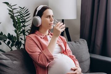 Cute young pregnant woman listening to music in wireless headphones, enjoy pure fresh cool mineral...