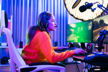 Young Caucasian woman professional gamer wearing yellow hoodie sits on a chair with a gaming table with pc, keyboard, monitor, microphone. Prepare for competition, cast gameplay or record a podcast. © artitwpd