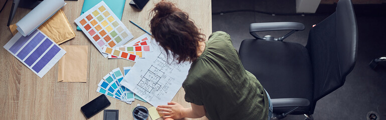 Top view of interior designer or architect in casual wear with messy hairdo working on a blueprint...