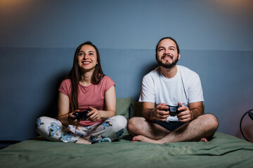 young latin couple gamers having fun playing video games on bed at night at home in Mexico Latin...