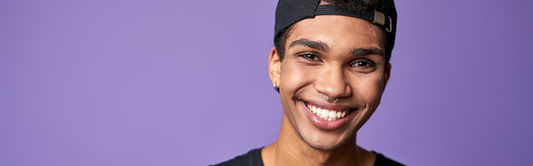 Portrait of smiling latino transgender man in black t-shirt and cap on purple background