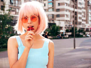 Young beautiful smiling hipster female in trendy summer clothes. Sexy carefree woman posing in the street in wig. Positive funny model having fun at sunset. In sunglasses. Eating candy lollipop