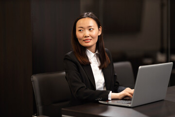 A beautiful Chinese woman is working on a laptop in her office. She is wearing formal clothes.