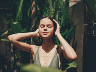 Fototapeta na wymiar A woman, a body in a swimsuit washes her head in a tropical shower outdoors against backdrop green tropical leaves, flowers and palm trees. Body and hair care, tanned skin, sunlight, smile, vintage