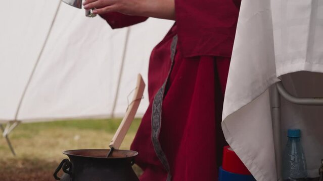 Female mag pours magic potion into small glass bottle. Woman makes drink in cauldron sitting at table near white tent. Activities of magician in medieval period