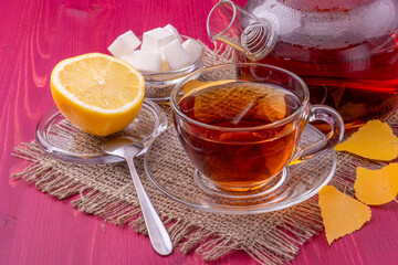 A teapot and a glass cup with brewing black tea. Next to the sliced lemon and lump sugar. Beautiful bright wooden background.