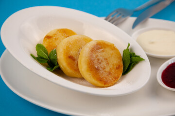 Round cottage cheese pancakes in a white plate on a blue background. Food breakfast. Healthy food. Sour cream sauce and mint. Top view closeup. Dessert meal. Curd cheese pancake ricotta fritters.