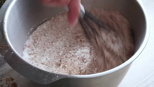 Top view of unrecognizable woman mixing ingredients: sugar, chocolate, flour with metal whisk in round metal bowl, preparing dough for baking cake. Confectionery, homemade dessert, pastry, cooking.