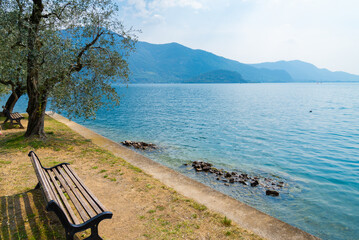 Fototapeta na wymiar A relaxing bench on the promenade of Monte Isola (Mount Island), Lake Iseo, Italy. Blue waters on the right, with italian alps and blue sky on the background.