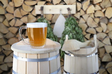 A glass mug of light beer against the background a stack of chopped birch firewood with hanging dry birch brooms and hat on it.
