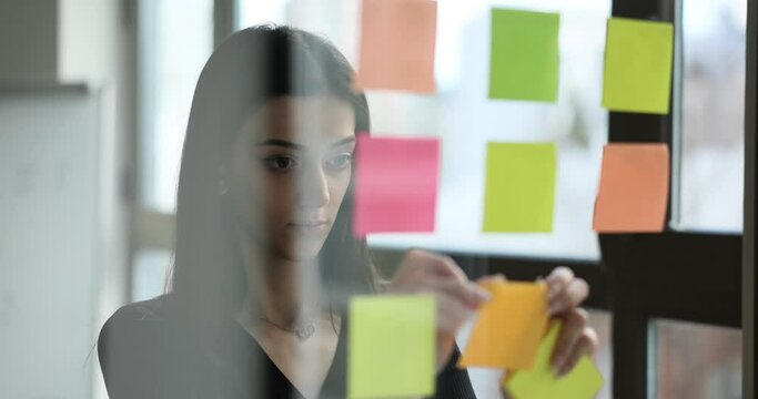 Business woman glues sticky papers with ready made tasks on glass. Manager posting notes successful coach leader planning corporate project near glass walls and adding ideas