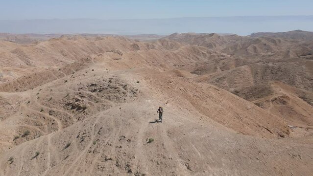 Man moutain biking downhill through a desert sharp mountain ridge fast with jumps, alone, during a sunny day with blue sky, aerial drone flying behind, near Arad mountains and the dead sea, Israel