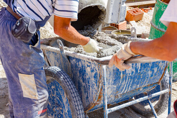 Team of workers are pouring concrete from cement mixer into wheelbarrow