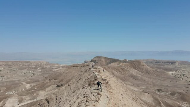 Drone passing through people with mountain bikes on a sharp ridge in the desert near Arad and the dead sea in Israel during a clear day with blue sky in winter with dramatic scenery and mountains