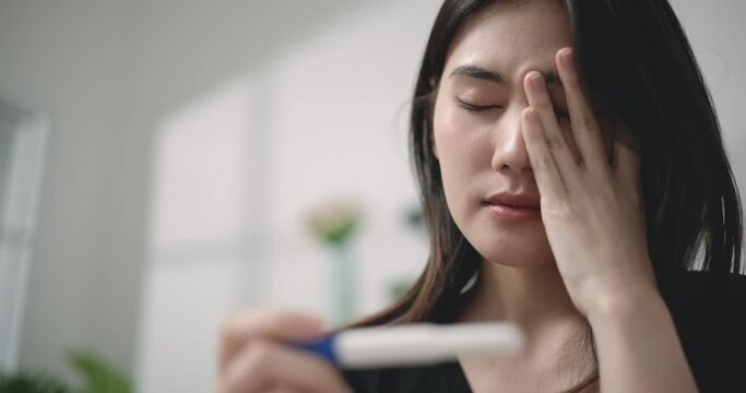 Footage close-up of Unhappy Asian young woman shocked at positive pregnancy test result while sitting on couch at the living room. Pregnancy or health problem concepts.