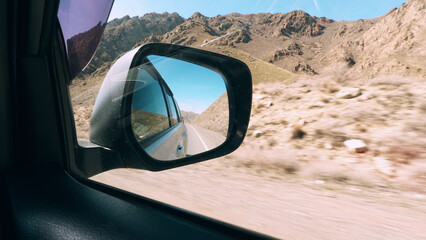 View of the road in the side mirror of the car. Behind there are yellow fences, huge rocky...
