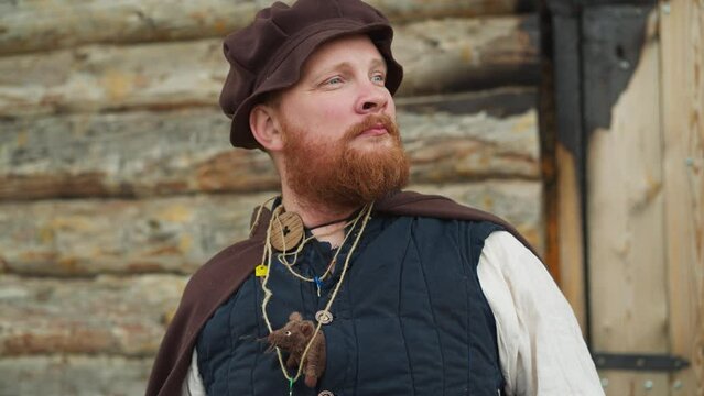 Bearded man in medieval clothes stands near wooden house. Knight with key on neck and mouse in bosom looks into distance dreamy closeup horizon tilts