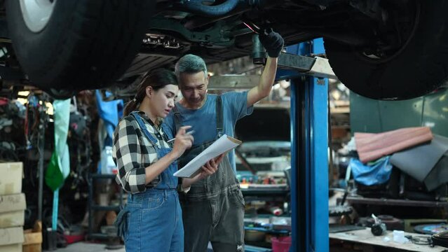 Asian automotive mechanic man and woman work in mechanics garage. Technician team of vehicle service manager worker look under car condition, Maintenance check to repair engine machine in car workshop