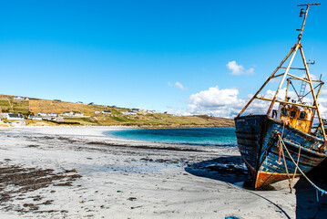 Fototapeta na wymiar Abandoned boat or fishing trawler on Arranmore island, Republic of Ireland. The sun is shining on a rusty vessel stranded in Aphort Strand, County Donegal. Forsaken slanting ship on Irish white beach