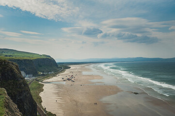 Aerial view of a dramatic cliff with the Downhill Beach and the imposing ocean underneath. Faraway cars parked on Downhill strand on a sunny day with small people walking on the huge shore by the sea
