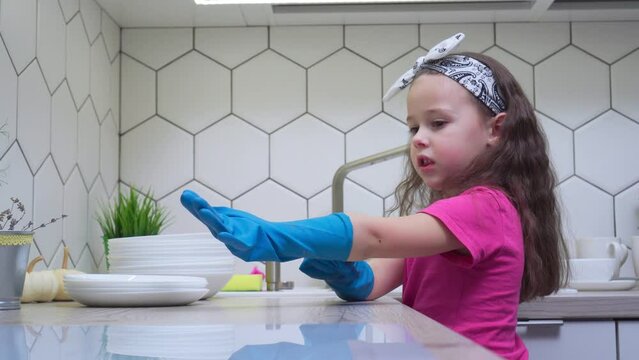 Side view of little preteen girl with loose long dark hair wearing pink T-shirt, putting on big blue rubber gloves for washing dishes near stack of clean white plates in kitchen. Housework, cleaning.