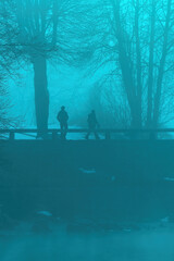 Two people walking over the old stone bridge at Bohinj lake in foggy winter morning