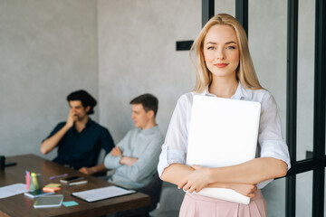 Portrait of friendly female entrepreneur in casual clothes holding laptop in hand standing in office, looking at camera. Startup business team discussing project sitting at desk on background.
