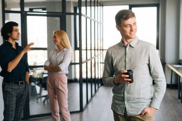 Fototapeta na wymiar Portrait of cheerful handsome male office worker in casual clothes holding takeaway coffee cup in hand standing in office, smiling looking away. Startup business team friendly chatting on background