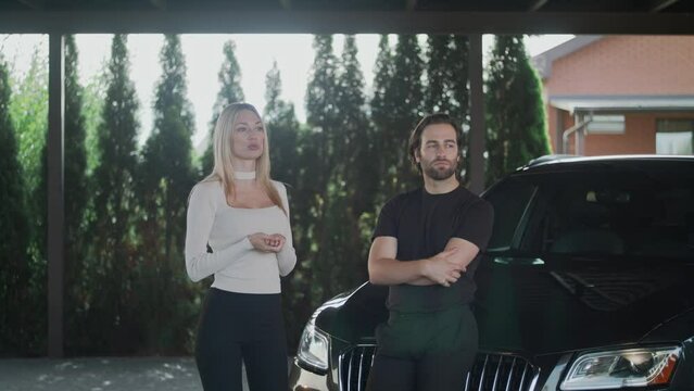 A couple is standing near their car. The girl isn't happy with her boyfriend's behavior. She gesticulates with her hands and shows her displeasure. The man is upset and doesn't want to listen to it.