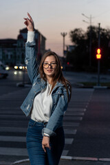 Fototapeta premium Portrait of an energetic girl in denim clothes on evening city background. Stylish young woman in glasses on city street